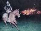 A person acting as the headless horseman riding a brown horse on a green lawn with a bonfire in the background